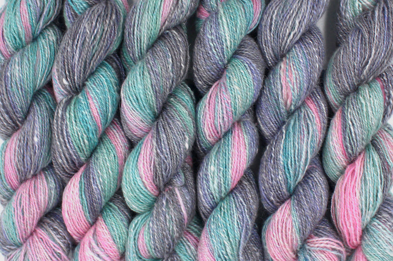 Six one of a kind, hand dyed gradient skein of multicolored Light Pink, Sky Blue, Seafoam Green, Grey, and Lavender self-striping wool Yarn lined up side by side.
