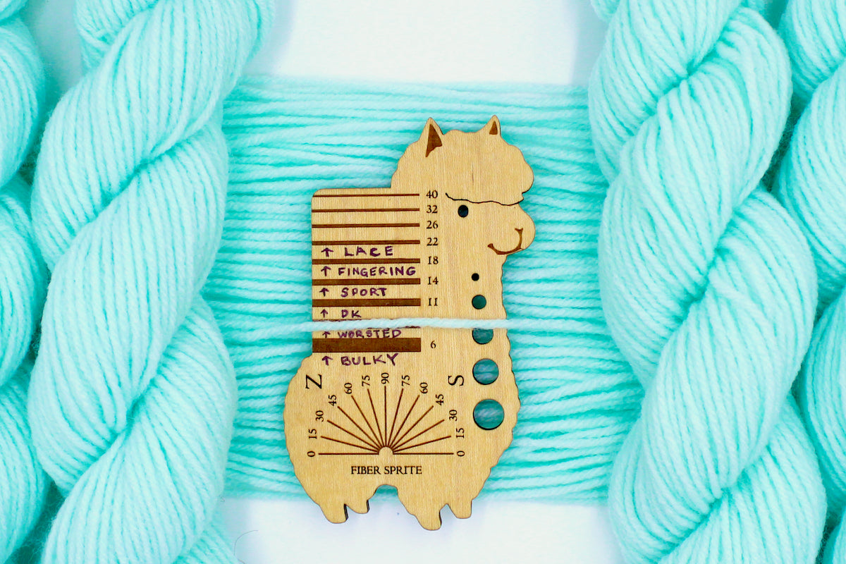 A skein of Vegan, Light Pastel Aquamarine 100% Acrylic, Worsted Weight Yarn recycled by hand from unwanted sweaters draped over the center of the frame with one strand on top of a Llama shaped Yarn weight tool, to better illustrate the thickness of the yarn.