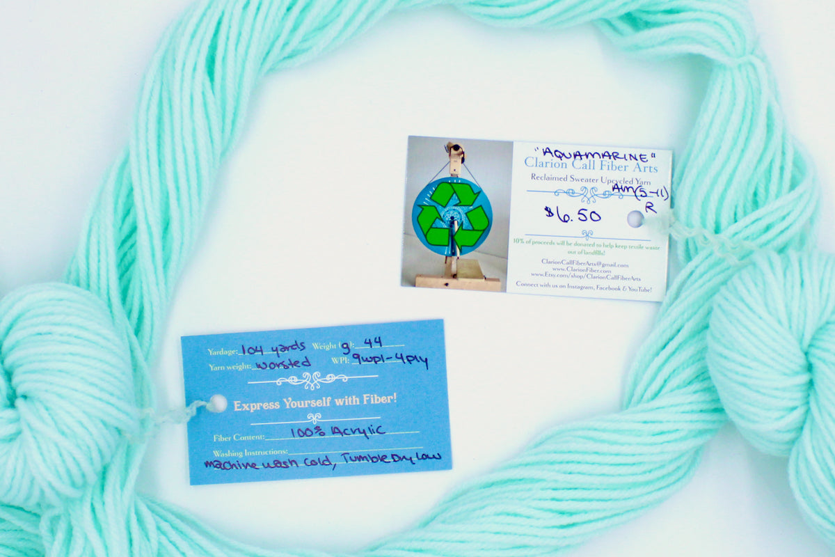 A skein of Vegan, Light Pastel Aquamarine 100% Acrylic, Worsted Weight yarn encircling the sweater labels it was recycled from, as well as the hand written yarn tags detailing the specs of this recycled yarn to help you make your next project!