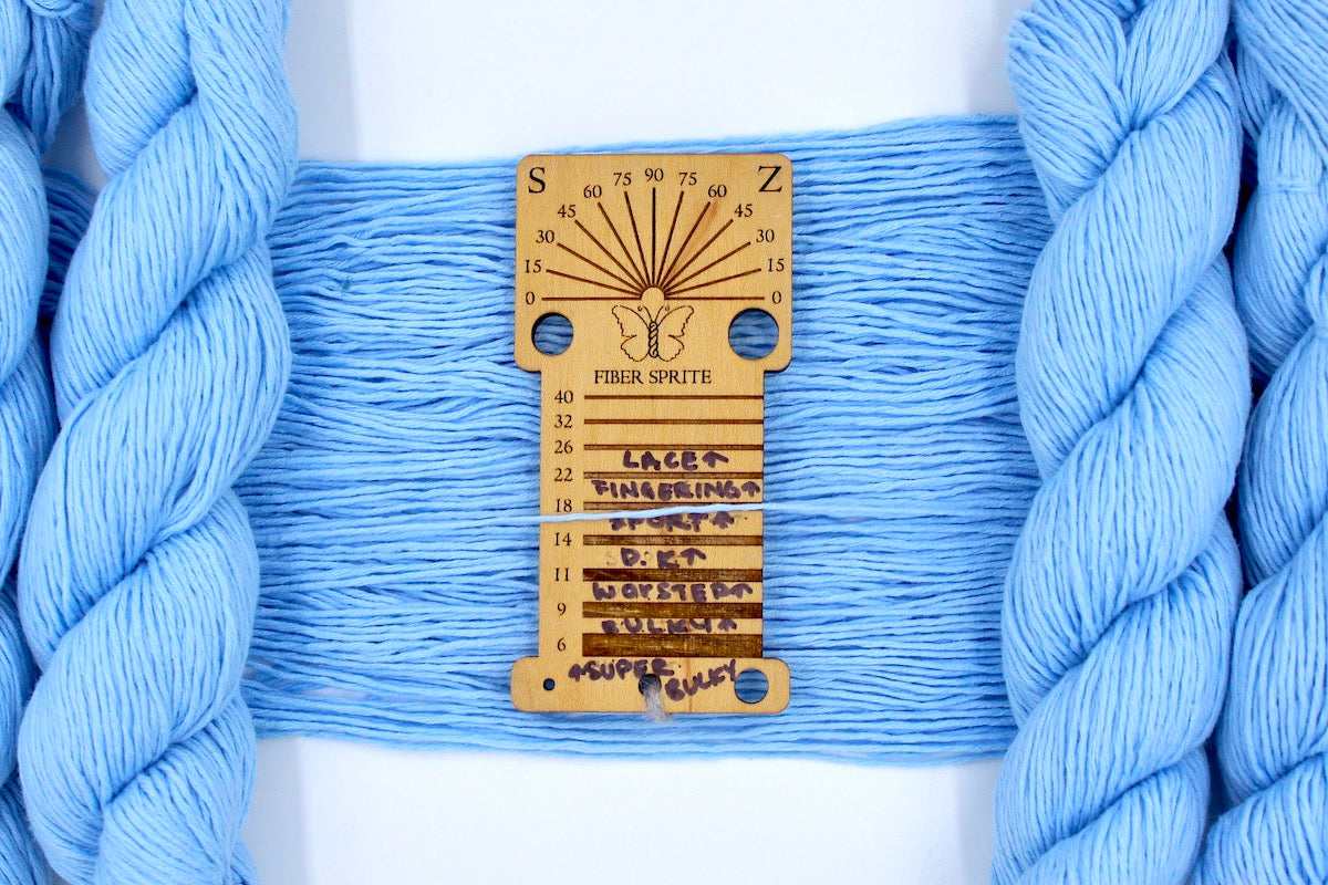 A skein of sport weight baby blue Yarn recycled by hand from unwanted sweaters draped over a Yarn weight tool