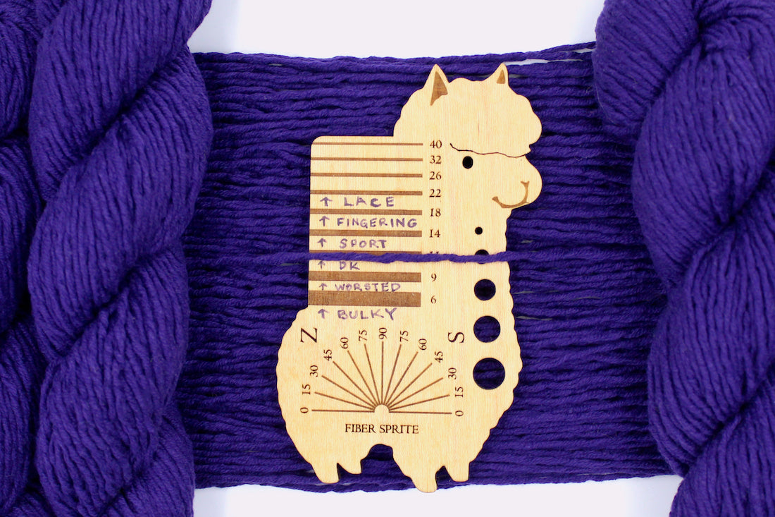 A skein of Vegan, Purple, 62% Cotton, 38% Acrylic, DK Weight Yarn recycled by hand from unwanted sweaters draped over the center of the frame with one strand on top of a Llama shaped Yarn weight tool, to better illustrate the thickness of the yarn.