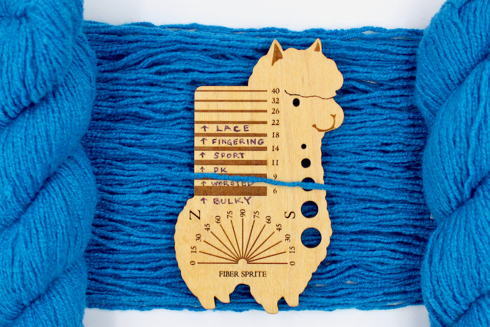 A skein of Bright Sky Blue, Wool, Acylic, Nylon, Angora, Worsted Weight Yarn recycled by hand from unwanted sweaters draped over the center of the frame with one strand on top of a Llama shaped Yarn weight tool, to better illustrate the thickness of the yarn.
