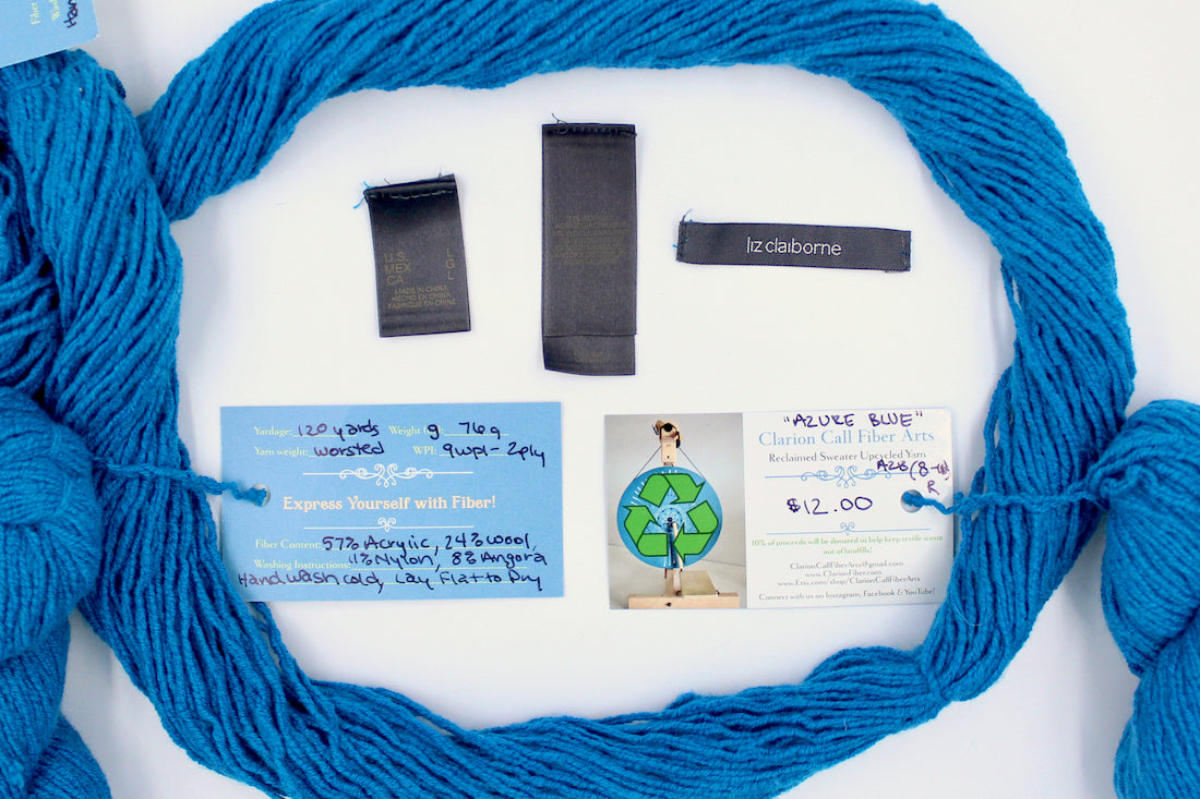 A skein of Bright Sky Blue, Wool, Acylic, Nylon, Angora, Worsted Weight yarn encircling the sweater labels it was recycled from, as well as the hand written yarn tags detailing the specs of this recycled yarn to help you make your next project!