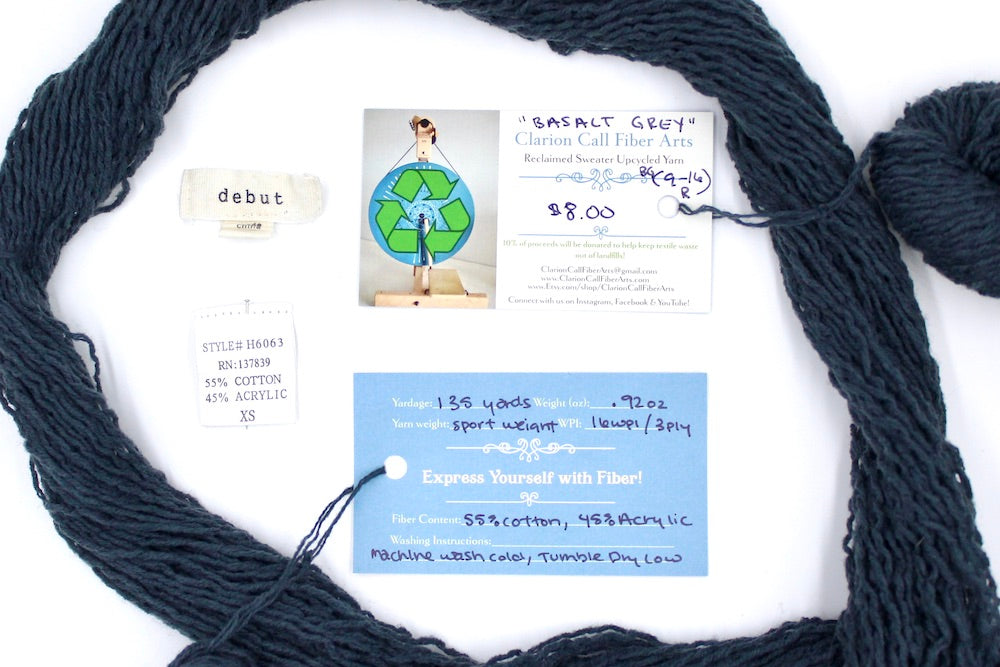 A skein of dark grey sport weight yarn encircling the sweater tags it was recycled from, as well as the hand written yarn tags detailing the specs of this upcycled yarn to help you make your next project!