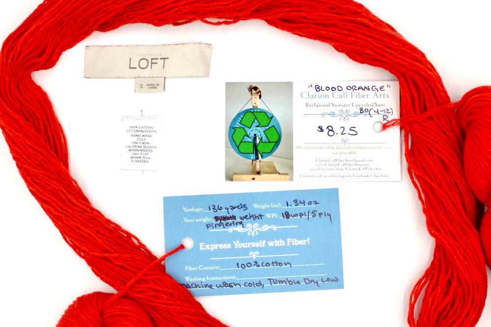 A skein of Bright Orange, Fingering weight yarn encircling the sweater tags it was recycled from, as well as the hand written yarn tags detailing the specs of this upcycled yarn to help you make your next project!