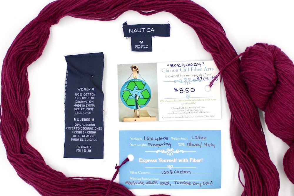 A skein of Maroon, Fingering weight yarn encircling the sweater tags it was recycled from, as well as the hand written yarn tags detailing the specs of this upcycled yarn to help you make your next project!