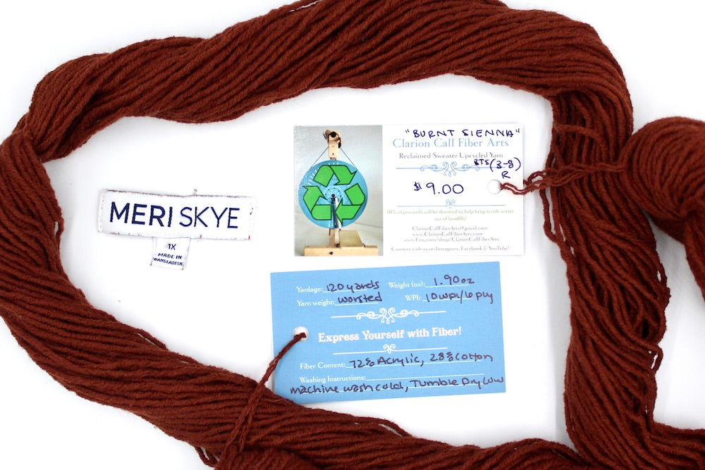 A skein of Reddish brown, Worsted weight yarn encircling the sweater labels it was recycled from, as well as the hand written yarn tags detailing the specs of this upcycled yarn to help you make your next project!