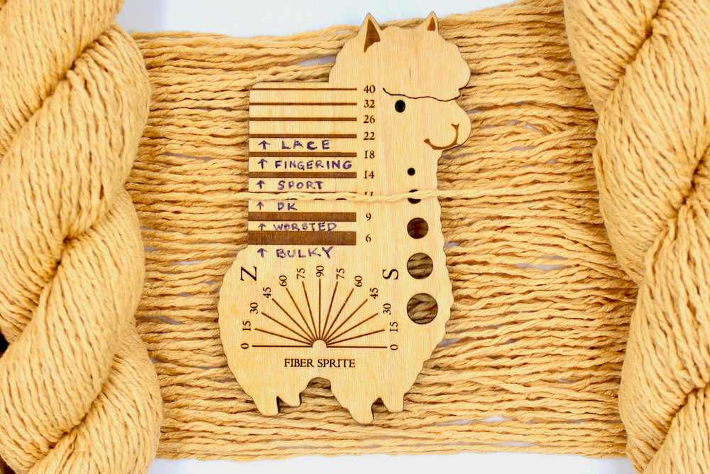 A skein of Vegan, Orangey Yellow, 60% Cotton, 40% Acrylic, DK Weight Yarn recycled by hand from unwanted sweaters draped over the center of the frame with one strand on top of a Llama shaped Yarn weight tool, to better illustrate the thickness of the yarn.