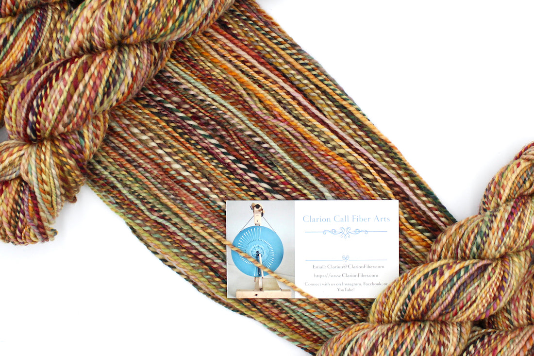 A one of a kind, DK Weight, hand dyed variegated skein of multicolored Orange, Rust, Green, and Gold, self-striping superwash wool Yarn draped diagonally across the frame, so you can really see the color play.