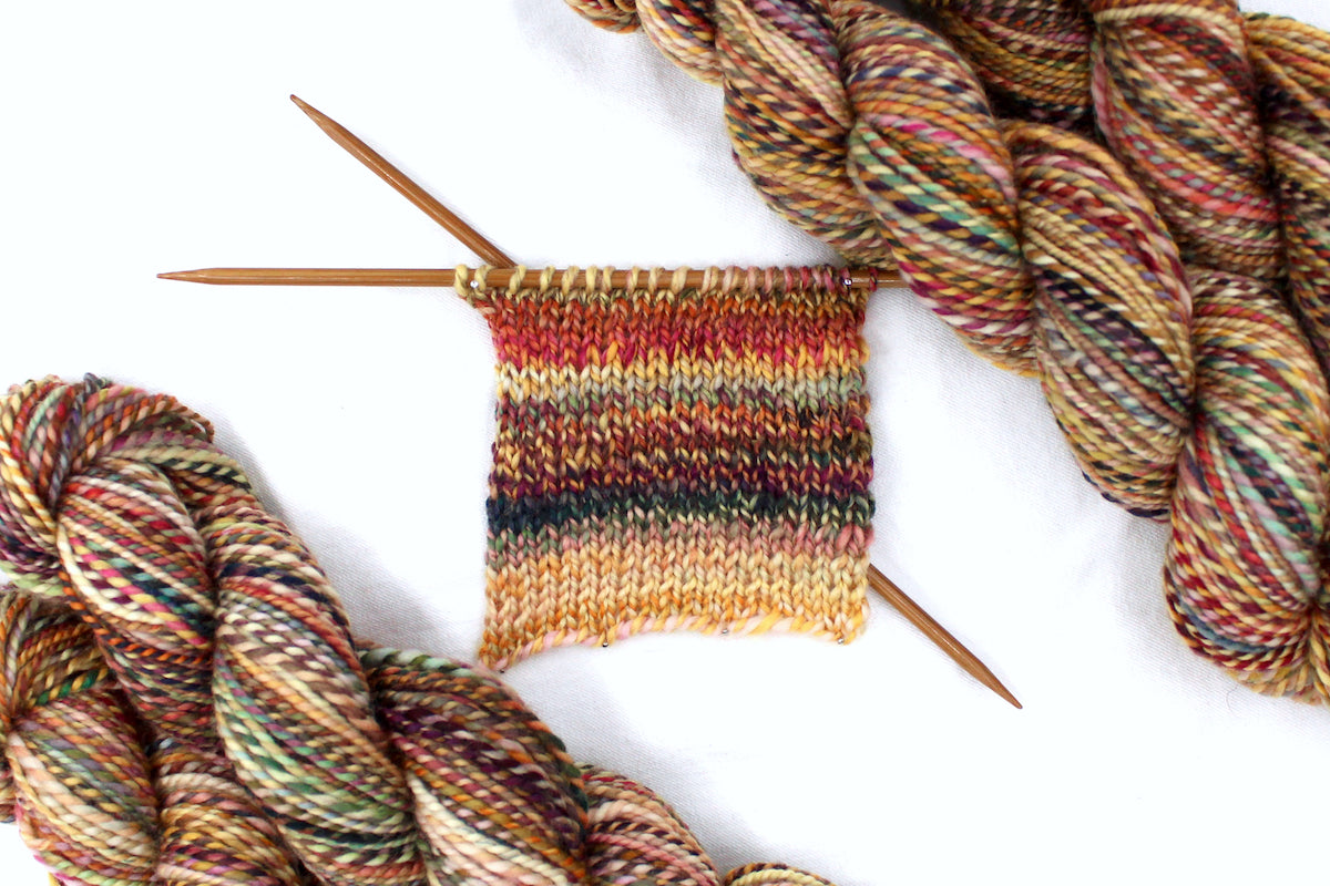 A sample swatch knitted from a one of a kind, DK Weight, hand dyed variegated skein of multicolored Orange, Rust, Green, and Gold, self-striping superwash wool Yarn.