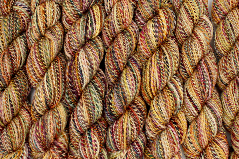 Six one of a kind, DK Weight, hand dyed variegated skein of multicolored Orange, Rust, Green, and Gold, self-striping superwash wool Yarn lined up side by side.