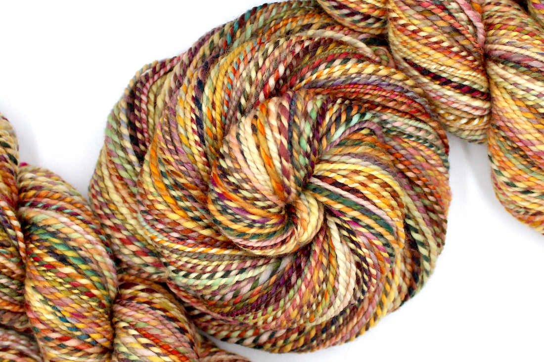 A one of a kind, DK Weight, hand dyed variegated skein of multicolored Orange, Rust, Green, and Gold self-striping superwash wool Yarn coiled attractively in the center of the frame.