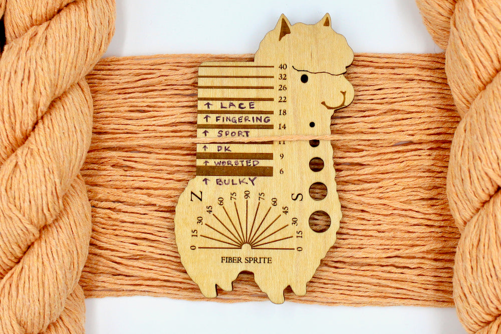 A skein of Vegan, Light Orange, 100% Cotton, DK Weight Yarn recycled by hand from unwanted sweaters draped over the center of the frame with one strand on top of a Llama shaped Yarn weight tool, to better illustrate the thickness of the yarn.