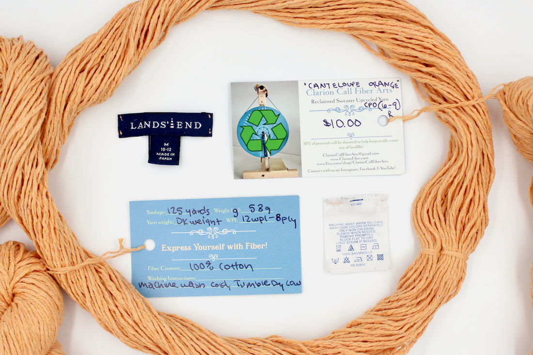 A skein of Vegan, Light Orange, 100% Cotton, DK Weight yarn encircling the sweater labels it was recycled from, as well as the hand written yarn tags detailing the specs of this recycled yarn to help you make your next project!