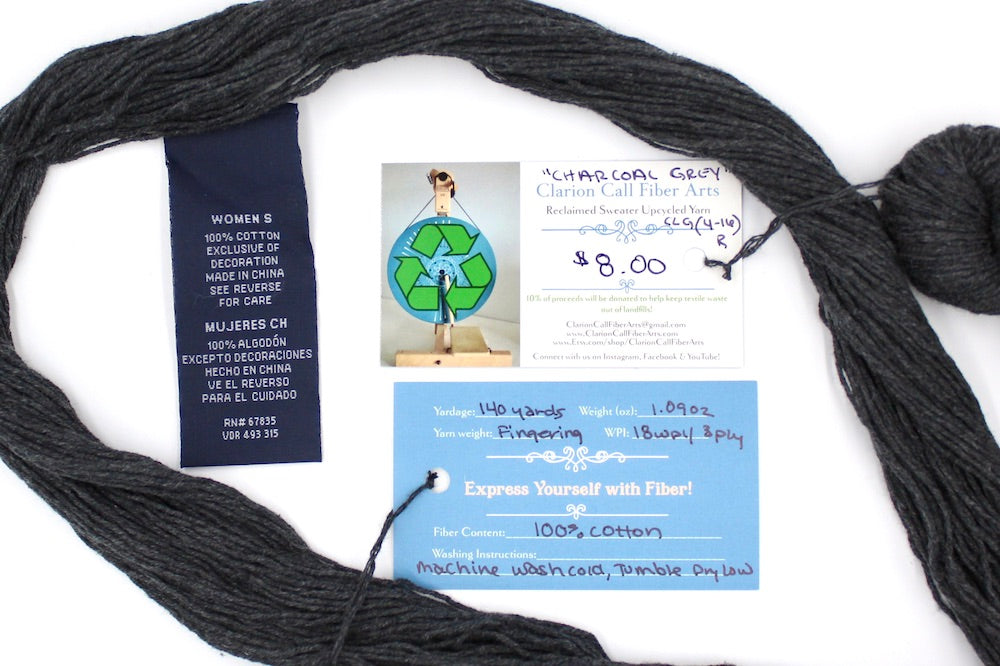 A skein of Dark Grey, Fingering weight yarn encircling the sweater labels it was recycled from, as well as the hand written yarn tags detailing the specs of this upcycled yarn to help you make your next project!