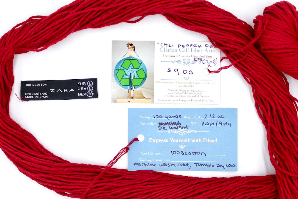 A skein of Ruby Red, Dk weight yarn encircling the sweater labels it was recycled from, as well as the hand written yarn tags detailing the specs of this upcycled yarn to help you make your next project!