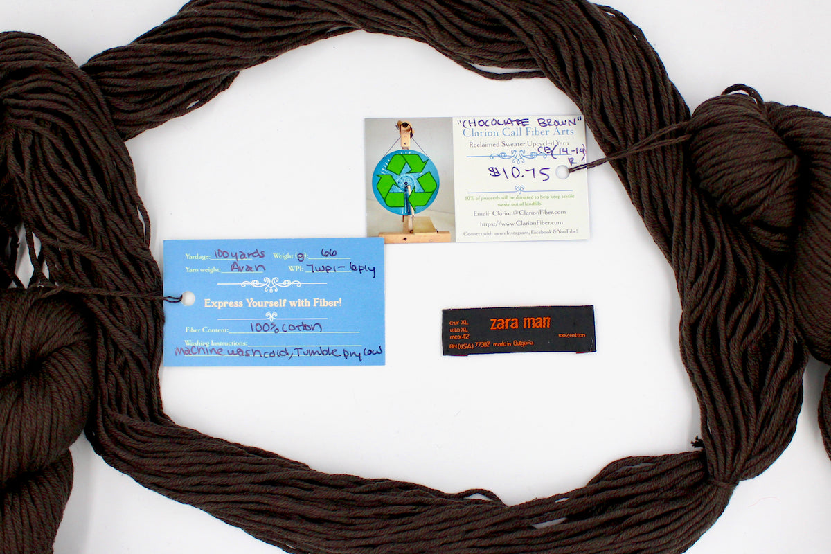 A skein of Vegan, Dark Chocolate Brown, 100% Upcycled Cotton, Heavy Worsted (or Aran) Weight yarn encircling the sweater labels it was recycled from, as well as the hand written yarn tags detailing the specs of this recycled yarn to help you make your next project!