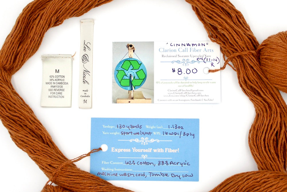 A skein of Brownish Orange, Sport yarn encircling the sweater labels it was recycled from, as well as the hand written yarn tags detailing the specs of this upcycled yarn to help you make your next project!