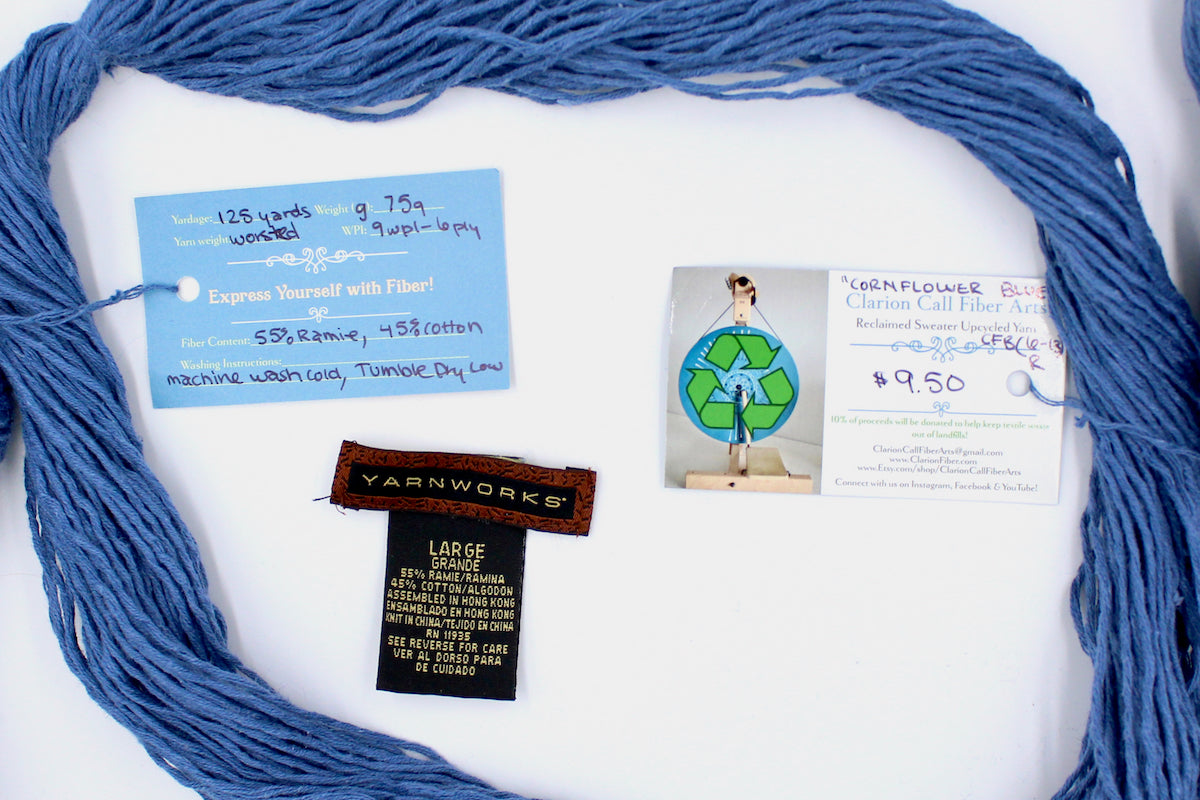 A skein of Vegan, Cornflower Blue, 55% Ramie, 45% Cotton, Worsted Weight yarn encircling the sweater labels it was recycled from, as well as the hand written yarn tags detailing the specs of this recycled yarn to help you make your next project!