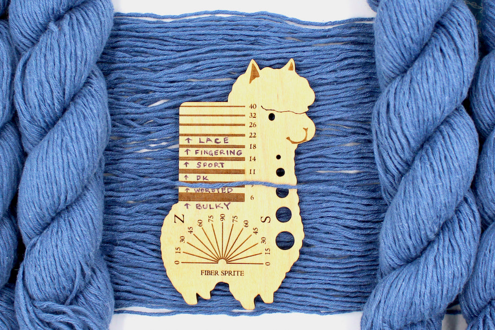 A skein of Vegan, Cornflower Blue, 55% Ramie, 45% Cotton, Worsted Weight Yarn recycled by hand from unwanted sweaters draped over the center of the frame with one strand on top of a Llama shaped Yarn weight tool, to better illustrate the thickness of the yarn.