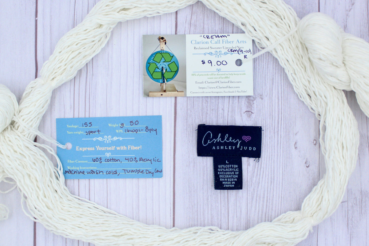 A skein of Vegan, Off-white, 60% Cotton, 40% Acrylic, Sport Weight yarn encircling the sweater labels it was recycled from, as well as the hand written yarn tags detailing the specs of this recycled yarn to help you make your next project!