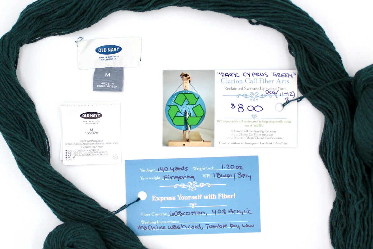 A skein of Dark Forest green, Fingering yarn encircling the sweater labels it was recycled from, as well as the hand written yarn tags detailing the specs of this upcycled yarn to help you make your next project!