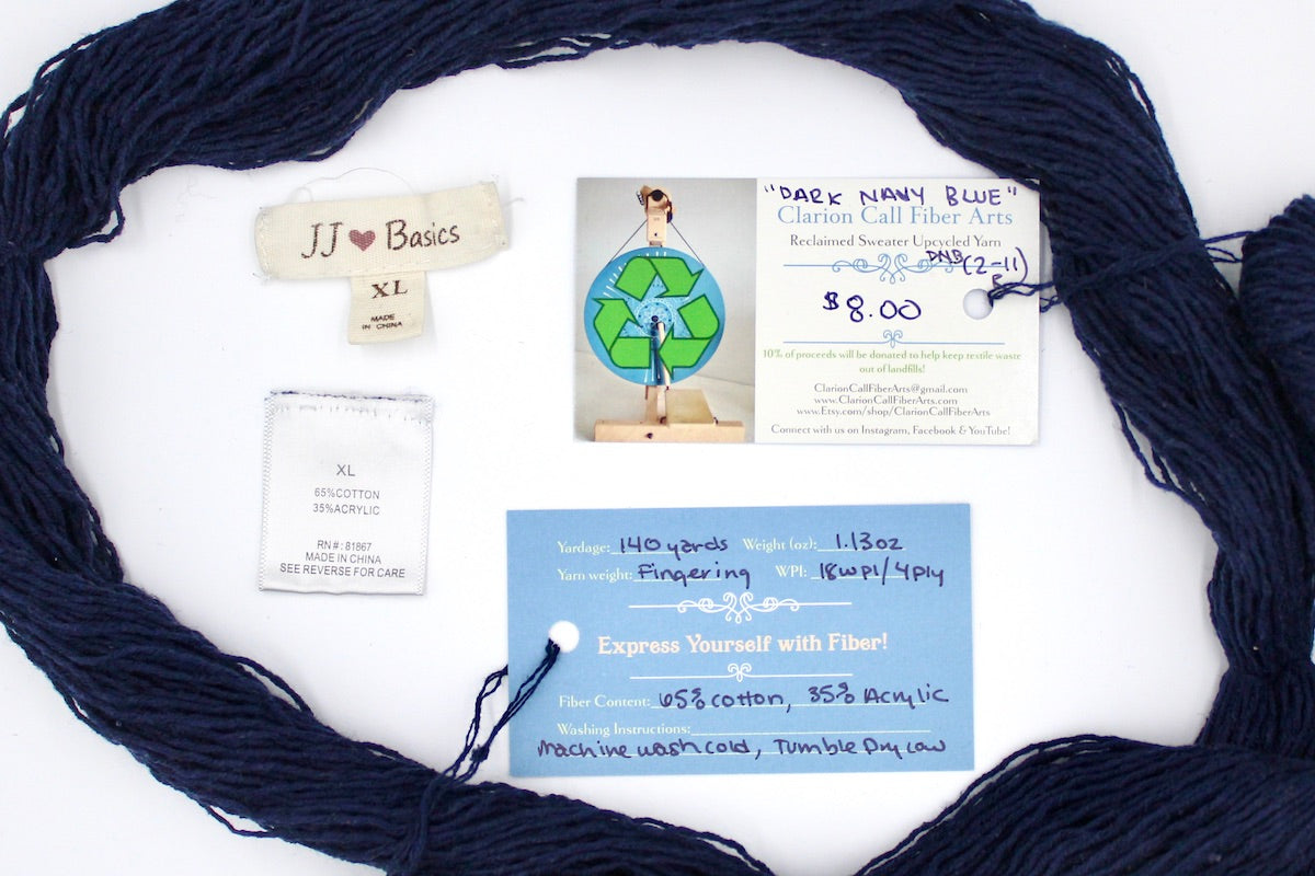 A skein of Dark Navy Blue, Fingering yarn encircling the sweater labels it was recycled from, as well as the hand written yarn tags detailing the specs of this upcycled yarn to help you make your next project!