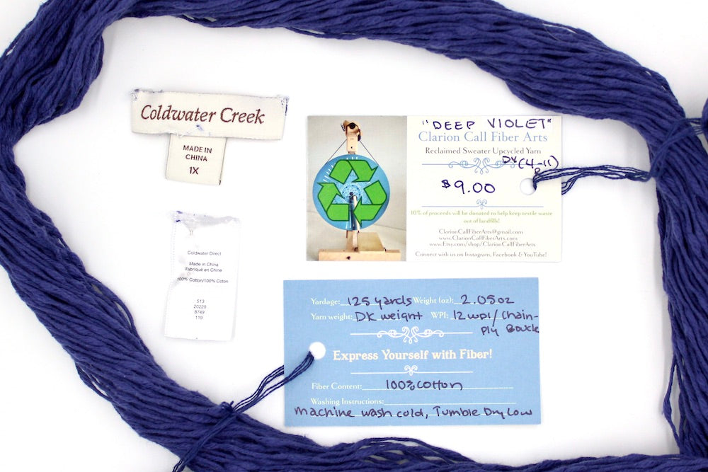 A skein of Deep Violet, Cotton Boucle, Dk Weight yarn encircling the sweater labels it was recycled from, as well as the hand written yarn tags detailing the specs of this upcycled yarn to help you make your next project!