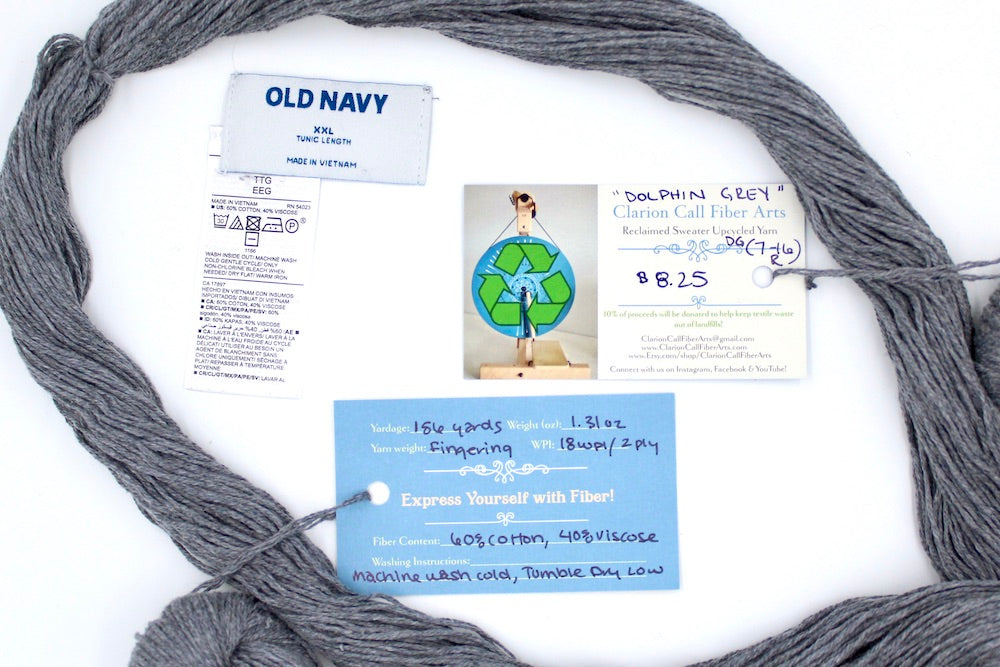 A skein of Light Grey, Cotton Viscose, Fingering Weight yarn encircling the sweater labels it was recycled from, as well as the hand written yarn tags detailing the specs of this upcycled yarn to help you make your next project!