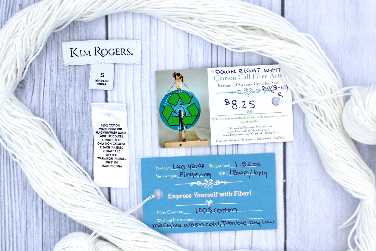 A skein of Pure White, Cotton, Fingering Weight yarn encircling the sweater labels it was recycled from, as well as the hand written yarn tags detailing the specs of this upcycled yarn to help you make your next project!