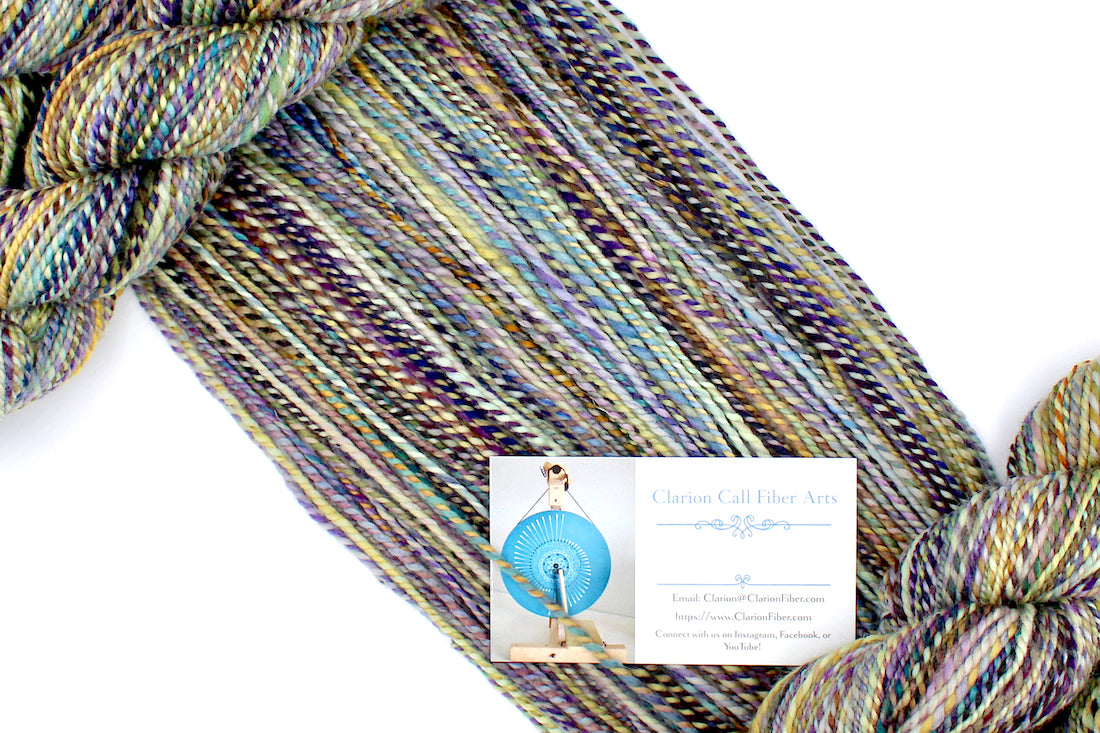 A one of a kind, Dk Weight, hand dyed variegated skein of multicolored Navy, Gold, and Purple self-striping 100% Superwash Merino wool Yarn draped diagonally across the frame, so you can really see the color play.