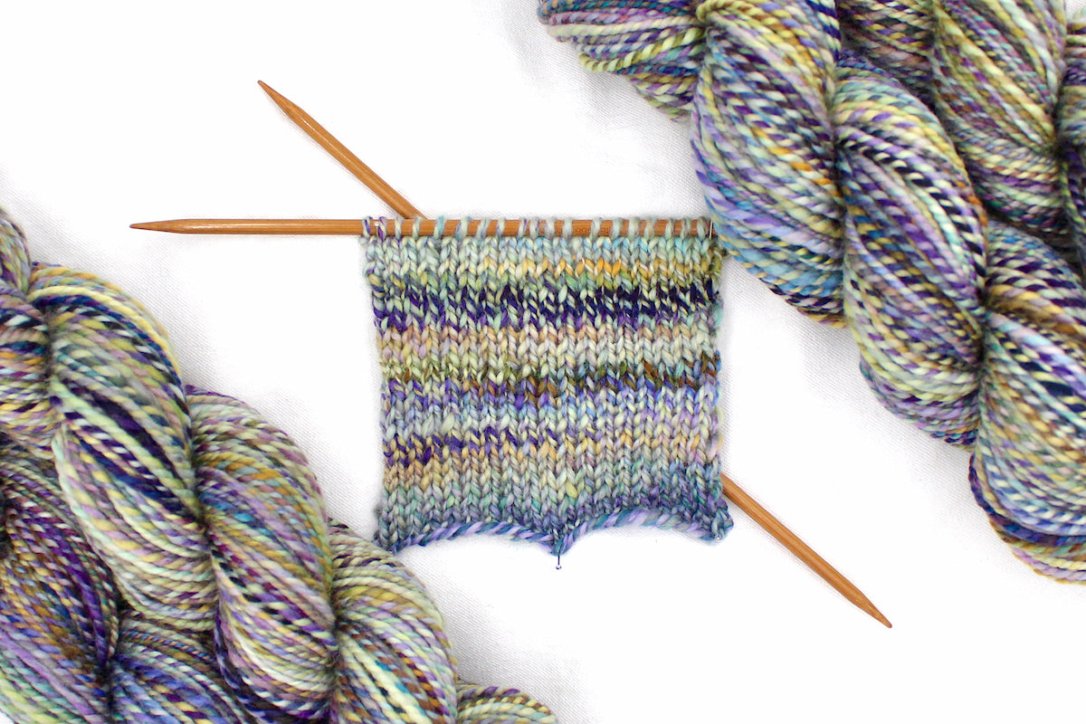 A sample swatch knitted on size 5 knitting needles from a one of a kind, Dk Weight, hand dyed variegated skein of multicolored Navy, Gold, and Purple self-striping 100% Superwash Merino wool Yarn.