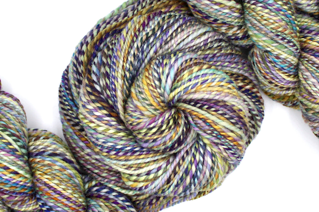 A one of a kind, Dk Weight, hand dyed variegated skein of multicolored Navy, Gold, and Purple self-striping 100% Superwash Merino wool Yarn coiled attractively in the center of the frame.