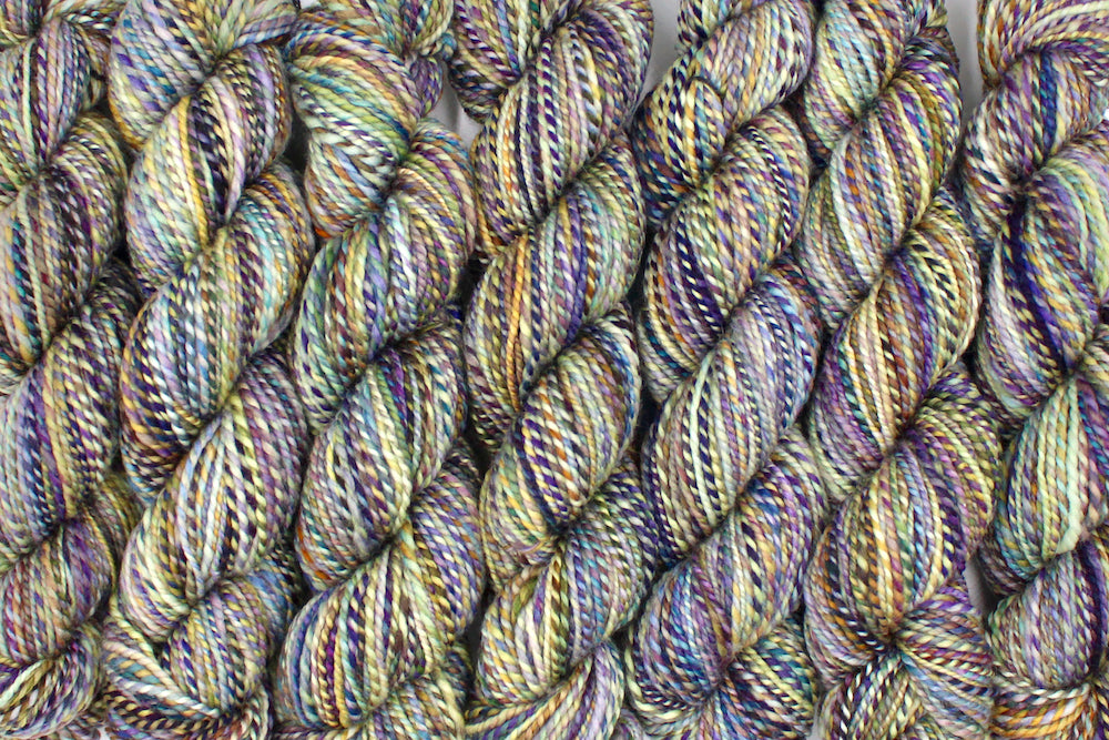 Six one of a kind, Dk Weight, hand dyed variegated skein of multicolored Navy, Gold, and Purple self-striping 100% Superwash Merino wool Yarn lined up side by side.