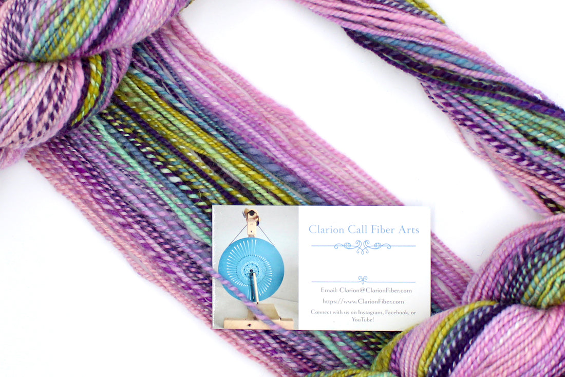 A one of a kind, Dk weight, hand dyed Gradient skein of multicolored Ballerina Pink, then move through Lavender, Magenta, Deep Purple, Baby Blue, Seafoam, and then finishes with Lime Green, Ombre self-striping Wool, and Mulberry Silk Yarn draped diagonally across the frame, so you can really see the color play.