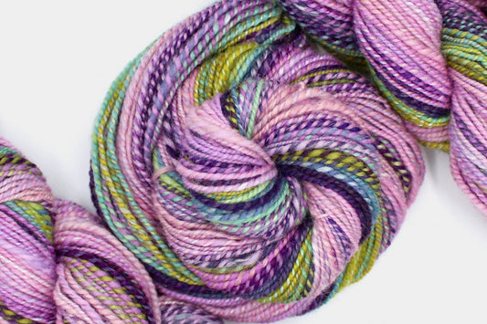 A one of a kind, Dk weight, hand dyed Gradient skein of multicolored Ballerina Pink, then move through Lavender, Magenta, Deep Purple, Baby Blue, Seafoam, and then finishes with Lime Green, Ombre self-striping Wool, and Mulberry Silk Yarn coiled attractively in the center of the frame.