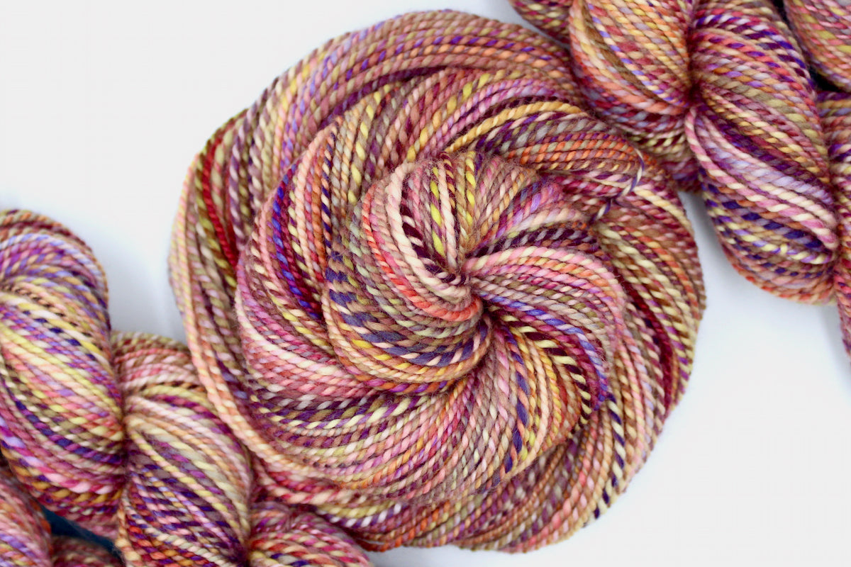 A one of a kind, hand dyed variegated skein of multicolored Orange, Pink, Yellow, and Purple self-striping wool Yarn coiled attractively in the center of the frame.