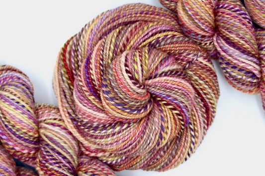 A one of a kind, hand dyed variegated skein of multicolored Orange, Pink, Yellow, and Purple self-striping wool Yarn coiled attractively in the center of the frame.