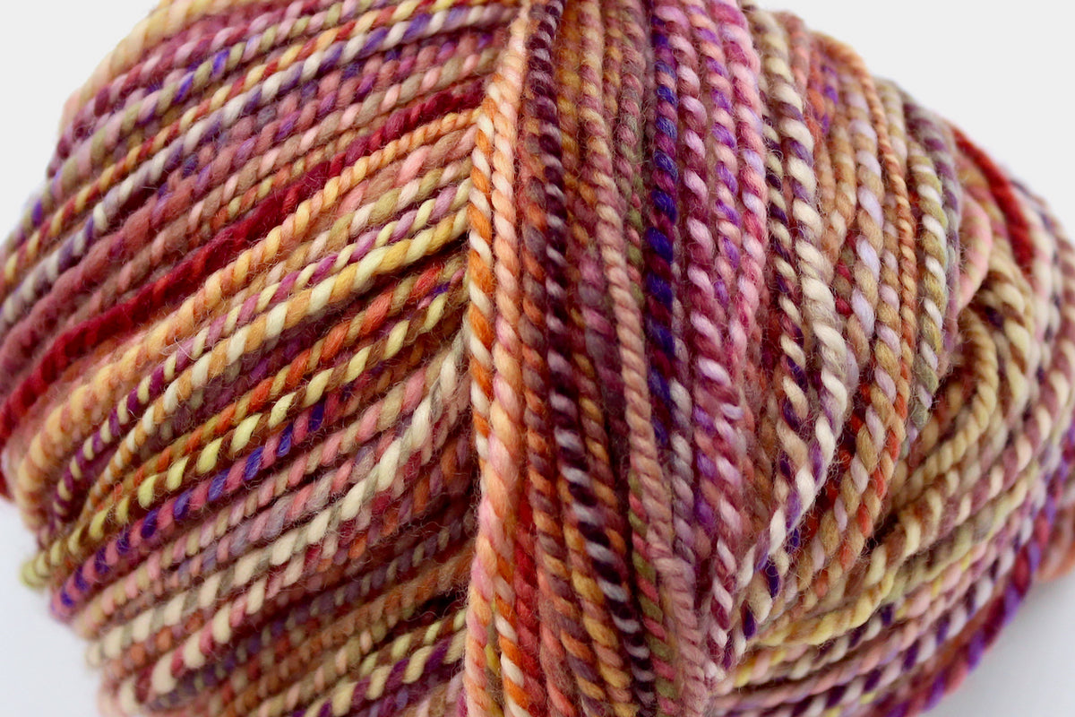A close up view of a one of a kind, hand dyed variegated skein of multicolored Orange, Pink, Yellow, and Purple self-striping wool Yarn.
