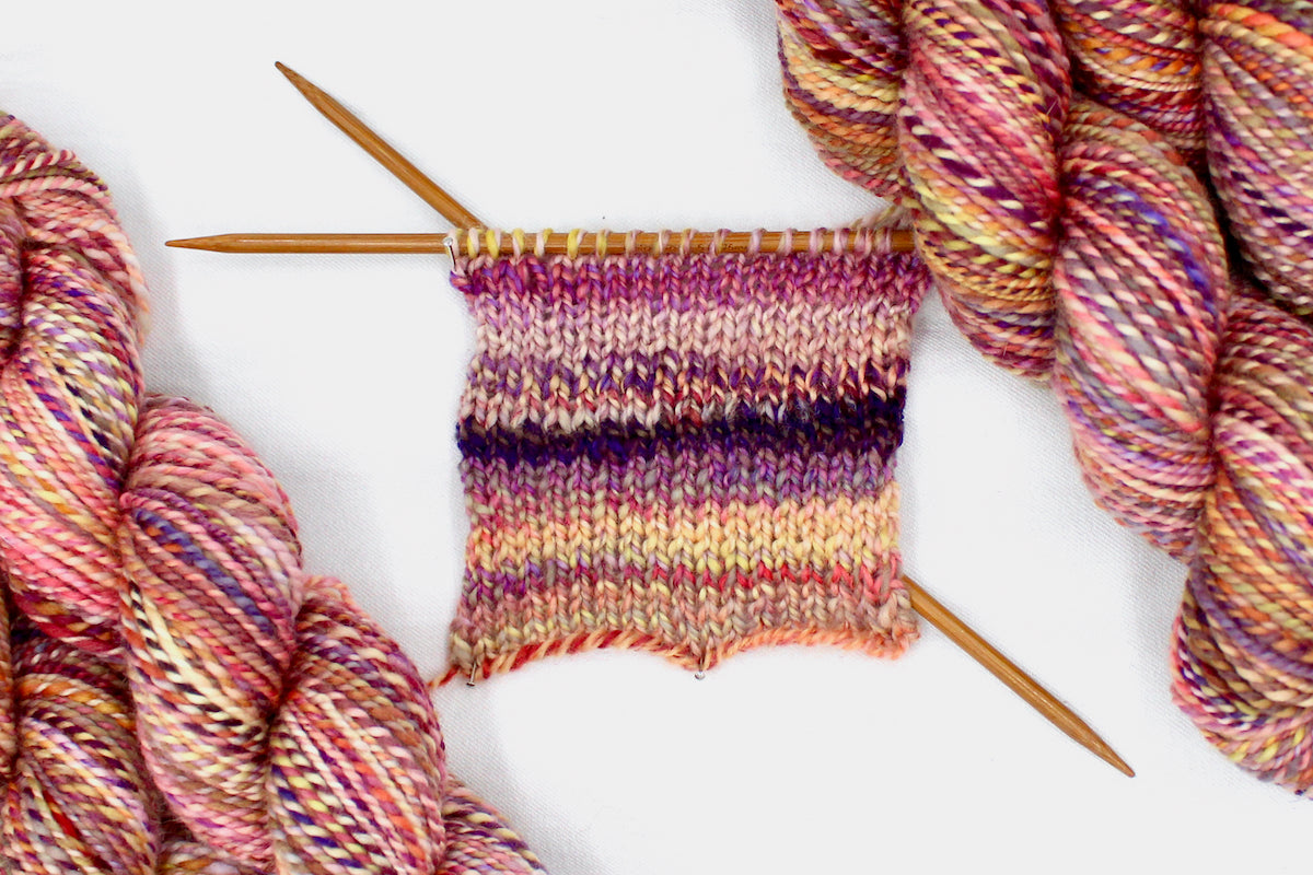 A sample swatch knitted from a one of a kind, hand dyed variegated skein of multicolored Orange, Pink, Yellow, and Purple self-striping wool Yarn.
