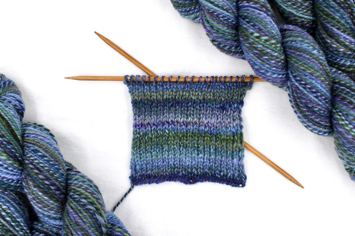 A sample swatch knitted from a one of a kind, Dk weight, hand dyed variegated skein of multicolored Purple, Lavender, Forest Green, Baby Blue, and Light Teal self-striping wool Yarn.