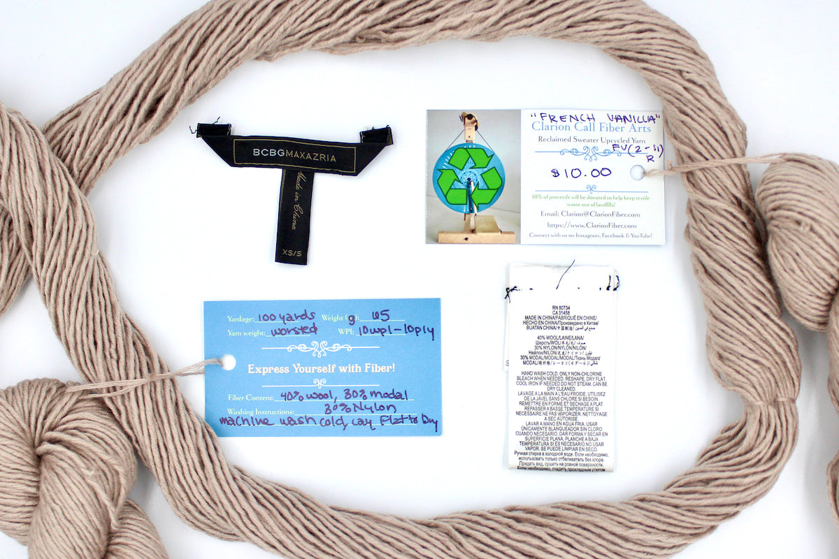 A skein of Tan, 40% Wool, 30% Nylon, 30% Modal, Worsted Weight yarn encircling the sweater labels it was recycled from, as well as the hand written yarn tags detailing the specs of this recycled yarn to help you make your next project!