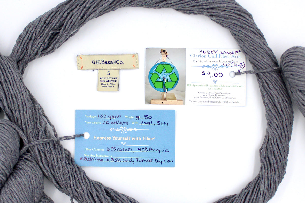 A skein of Vegan, Medium Grey, 60% Cotton, 40% Acrylic, Dk Weight yarn encircling the sweater labels it was recycled from, as well as the hand written yarn tags detailing the specs of this recycled yarn to help you make your next project!