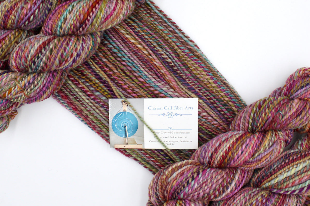 A one of a kind, Worsted weight, hand dyed variegated skein of multicolored Pink, Orange, Gold, Blue, Green, and Purple self-striping wool Yarn draped diagonally across the frame, so you can really see the color play.