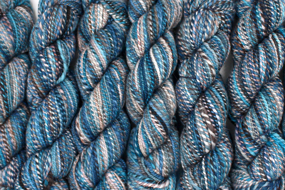 Six one of a kind, Dk Weight, hand dyed variegated skeins of multicolored Blue, Teal, Brown, and Taupe self-striping Hand Spun Yarn, made of 100% Hand Dyed Merino Wool lined up side by side.