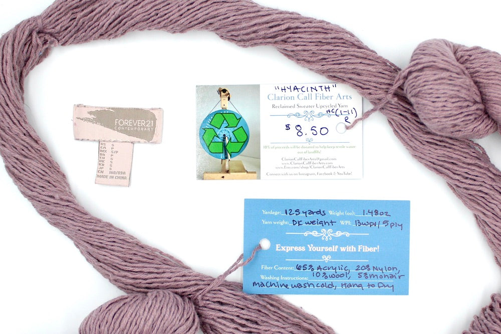 A skein of Dusky Mauve, Acrylic, Nylon, Wool, Mohair, Dk Weight yarn encircling the sweater labels it was recycled from, as well as the hand written yarn tags detailing the specs of this upcycled yarn to help you make your next project!