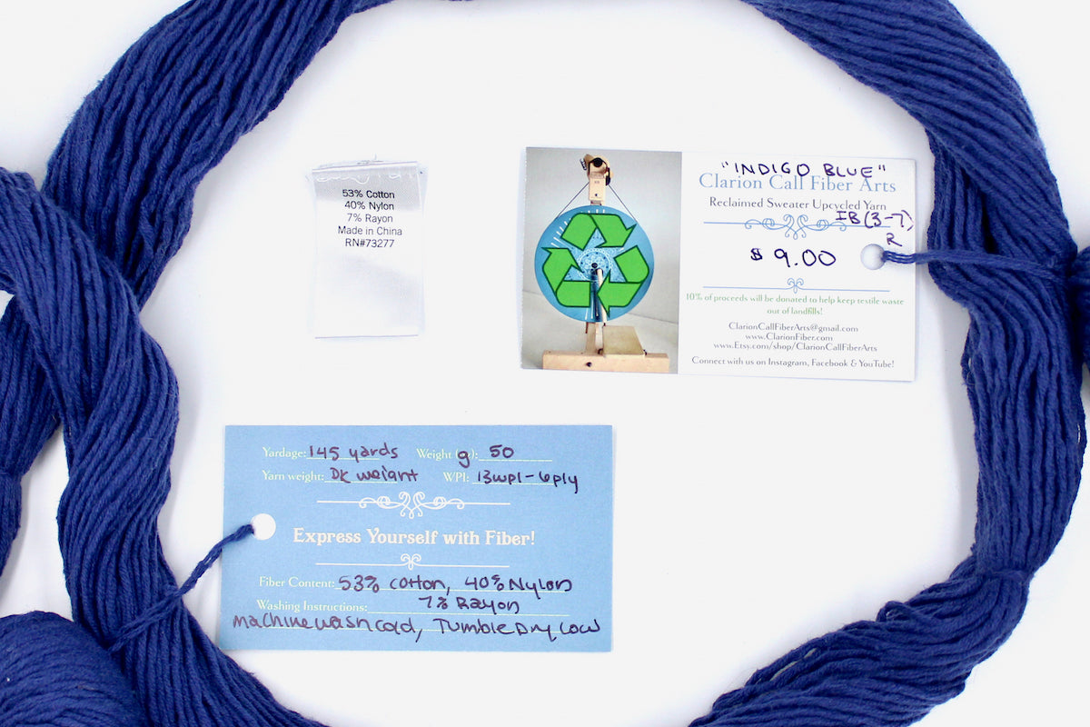 A skein of Vegan, Purplish Blue, Cotton, Nylon, Rayon, Dk Weight yarn encircling the sweater labels it was recycled from, as well as the hand written yarn tags detailing the specs of this recycled yarn to help you make your next project!