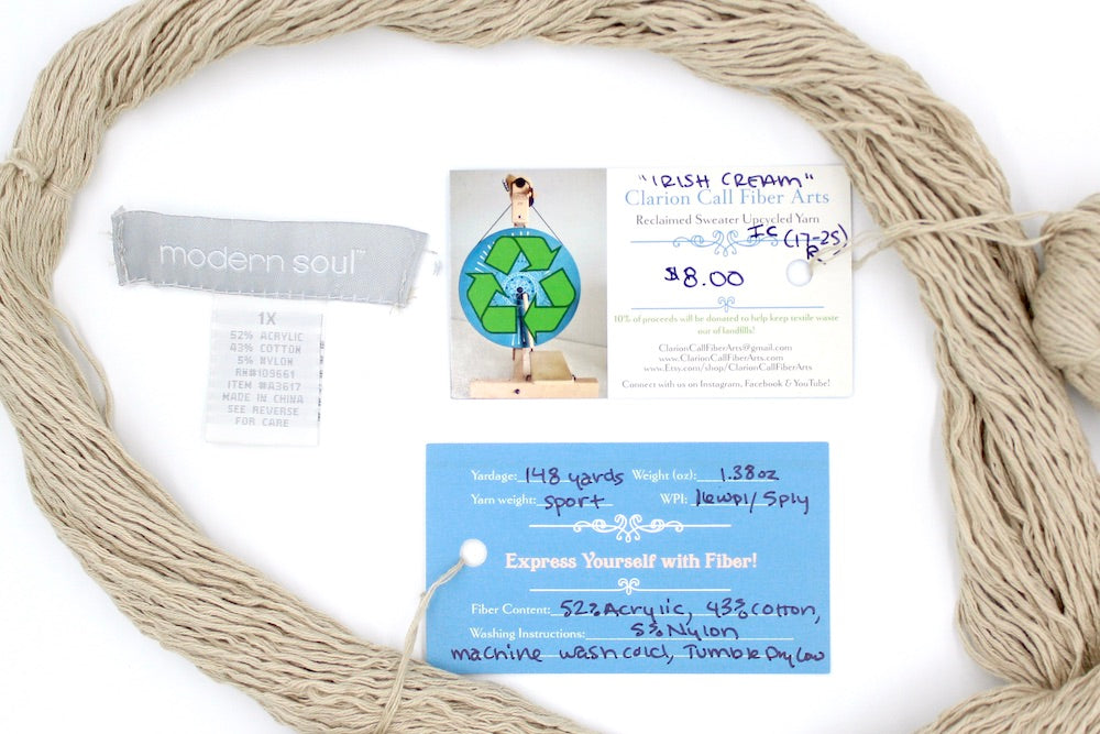 A skein of Vegan, Tan/ Beige/ Cream, Acrylic/ Cotton/ Nylon, Sport Weight yarn encircling the sweater labels it was recycled from, as well as the hand written yarn tags detailing the specs of this upcycled yarn to help you make your next project!
