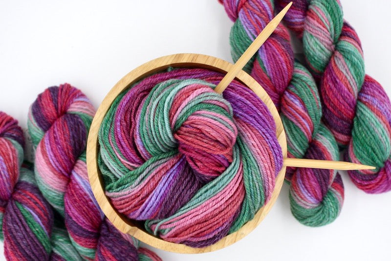 A one of a kind, hand dyed gradient skein of multicolored Plum Purple, Red, Fuchsia, Lavender, Pink, and Green self-striping wool Yarn, in a yarn bowl with knitting needles, ready to be made into something beautiful!