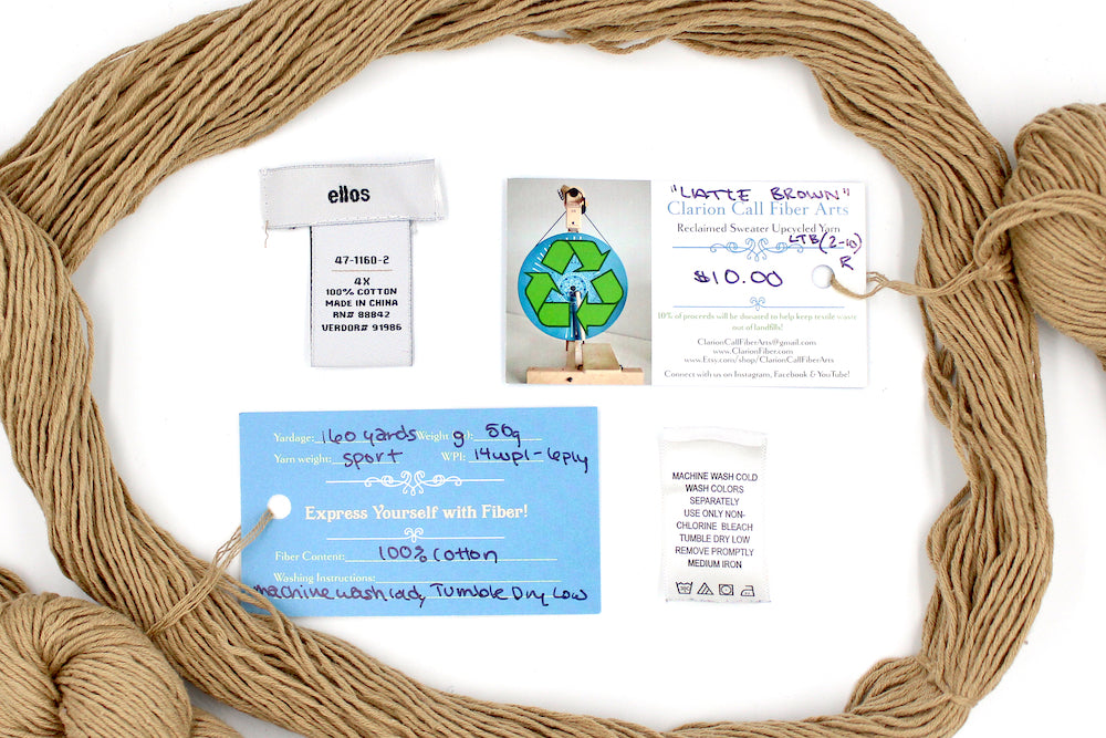 A skein of Vegan, golden tan, 100% Cotton, Sport Weight yarn encircling the sweater labels it was recycled from, as well as the hand written yarn tags detailing the specs of this recycled yarn to help you make your next project!